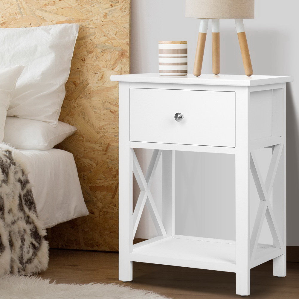 White nightstand with a drawer next to a bed with white pillows and a wooden headboard.