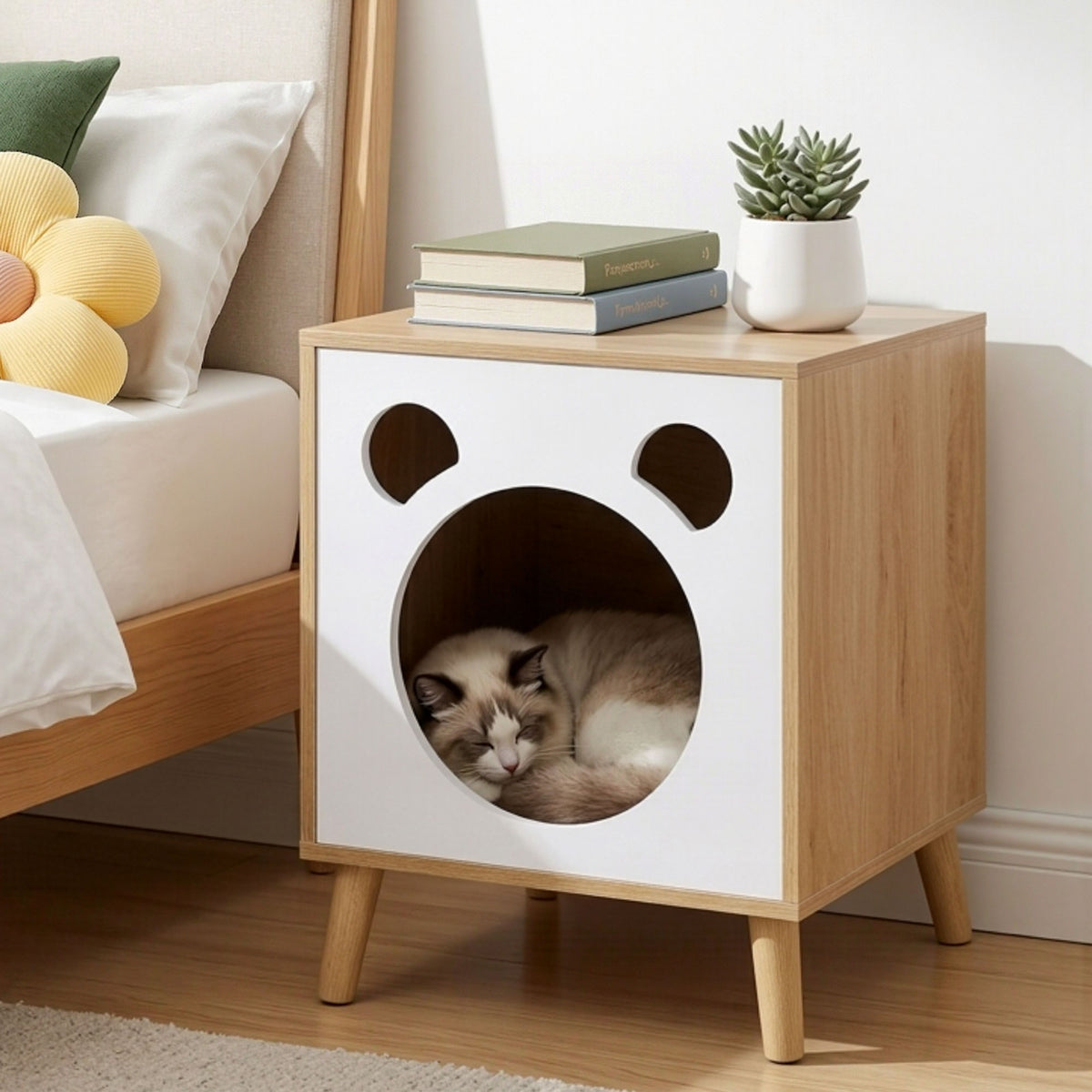Wooden pet bed with a cat inside, placed next to a sofa with cushions and books.