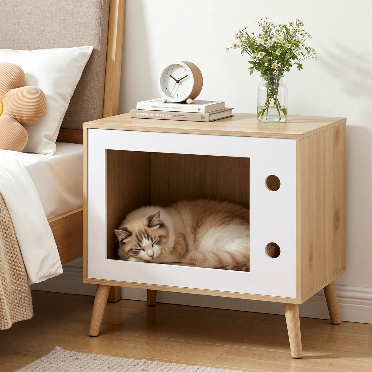 Cat lying inside a wooden and white pet bed next to a bed with pillows and a clock.