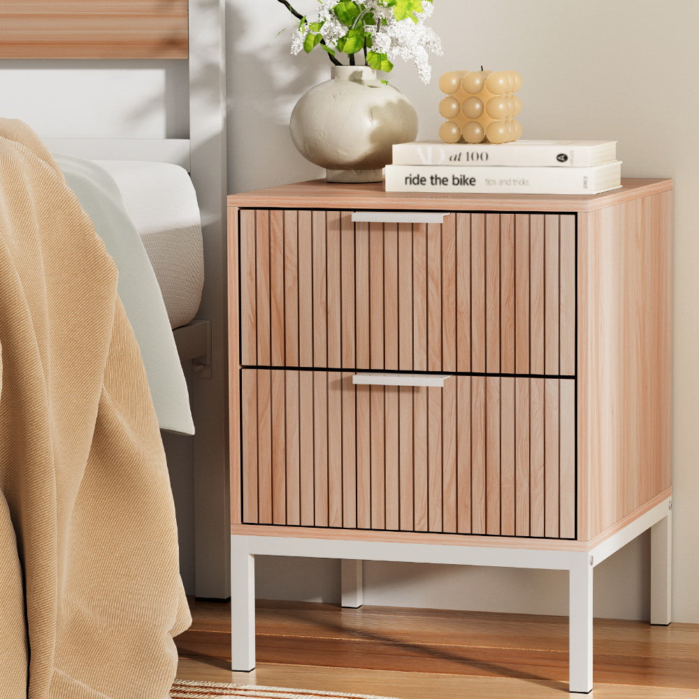 Wooden nightstand with two drawers, books, and decorative items next to a bed.