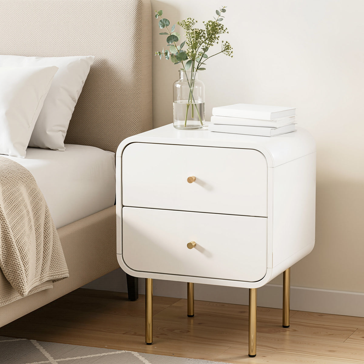 White nightstand with gold legs next to a bed with white pillows and a beige blanket.