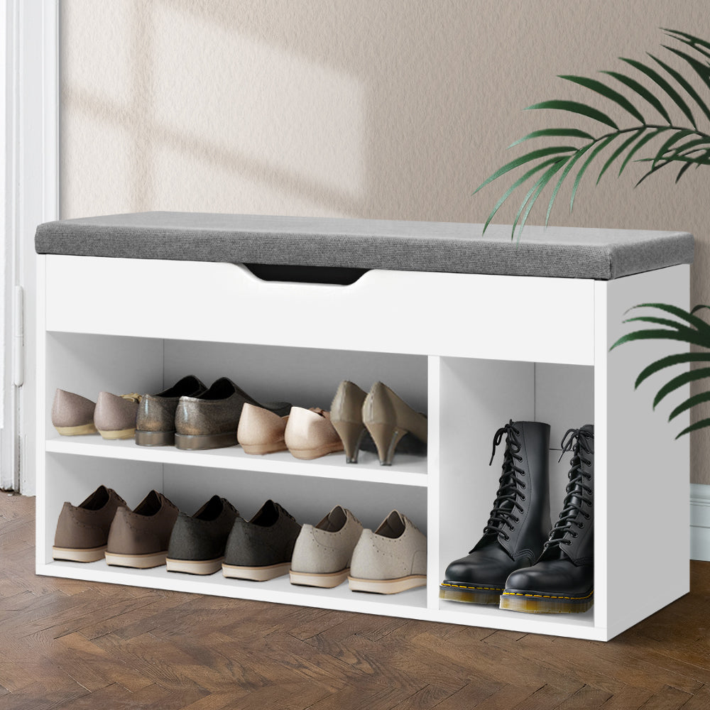 White shoe storage bench with gray cushion holding various shoes in a room with a plant.