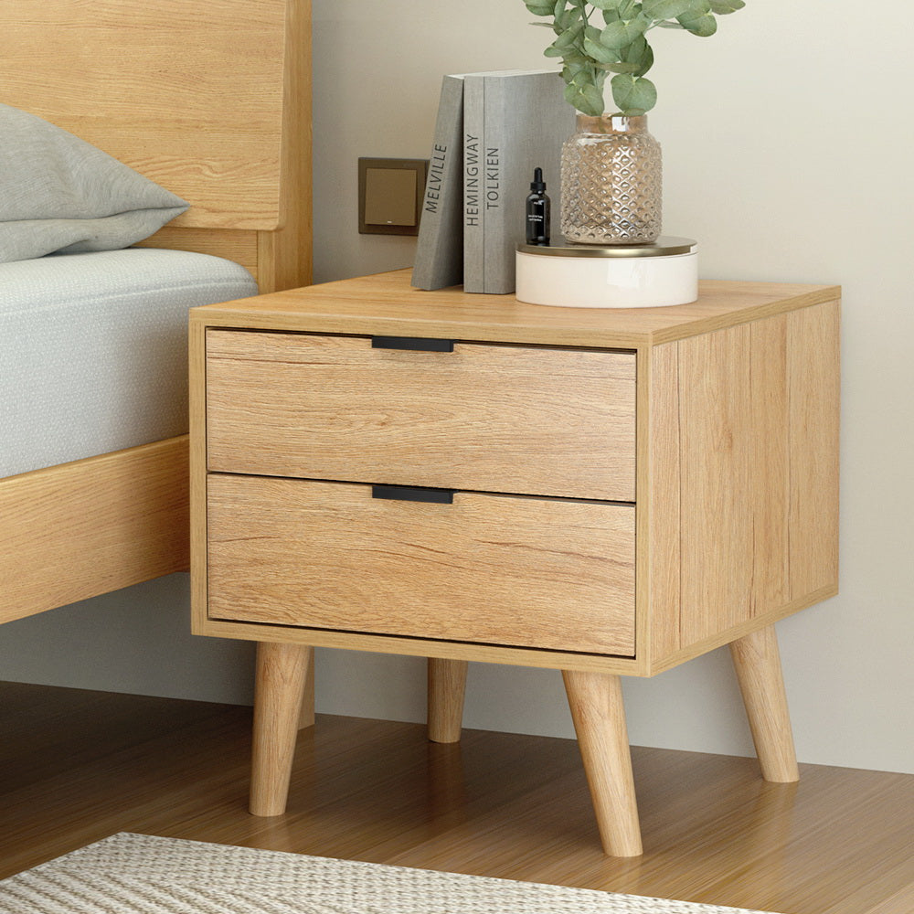 Wooden nightstand with two drawers next to a bed, featuring books and a plant.