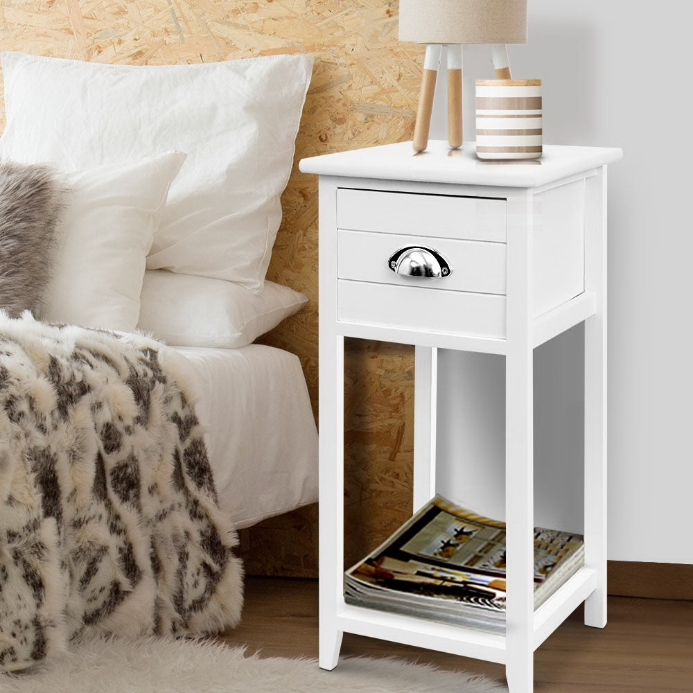 White nightstand with a lamp and books next to a bed with white pillows and a fur blanket.