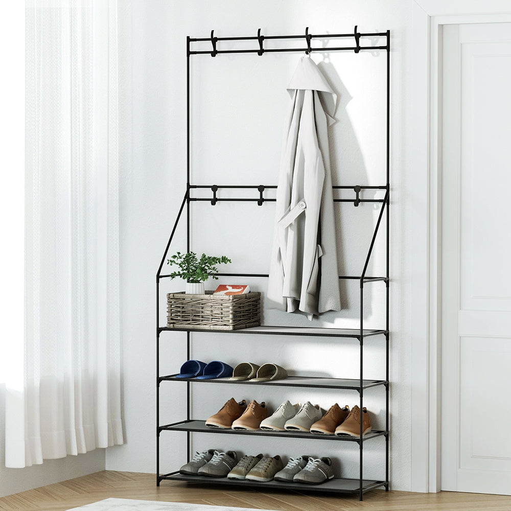 Black metal clothing rack with shelves holding shoes, a plant, and a coat against a white wall.